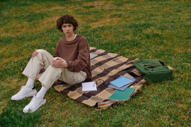 Genç bir adam çimenli bir alanda battaniyeye uzanıyor, doğanın ortasında sessiz bir yansımayla meşgul oluyor..