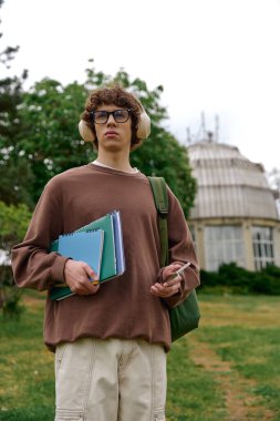 Genç adam dışarıda geziniyor, elinde defterler ve huzurlu bir ortamda bir telefon tutuyor..