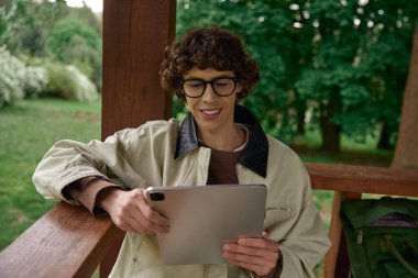 Etrafı yeşilliklerle çevrili ahşap bir verandada tablet kullanırken gülümseyen rahat genç adam..
