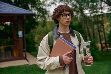 Yakışıklı genç bir öğrenci, elinde defterleri ve bir şişe suyla doğada geziniyor..