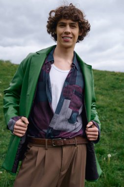 Yeşil ceketli ve sıradan giyinmiş genç bir adam çimenli bir alanda mutlu bir şekilde yürüyor..