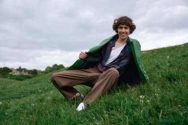 Sıradan giyinmiş genç bir adam çimenlerin üzerinde oturur, gülümserken, dışarıda huzurlu bir anın tadını çıkarır..