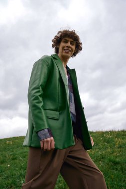 Neşeli genç bir adam çimenli bir alanda yürüyor, gülümsüyor ve şık kıyafetini sergiliyor..