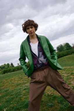 Yakışıklı genç adam yeşil bir parkta yürüyor, parlak bir ceket ve rahat bir pantolon giyiyor..