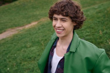 Canlı yeşil bir ceketle giyinmiş genç bir adam yemyeşil çimenlikte dolaşıyor, gülümsüyor ve rahatlıyor..