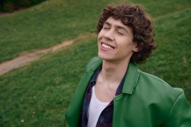 Genç ve yakışıklı bir öğrenci açık havada neşeli bir şekilde yürüyor, yemyeşil çimlerin arasında gülümsüyor..