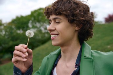 Yakışıklı genç adam elinde bir karahindiba tutarak doğada yürürken neşeyle gülümsüyor..