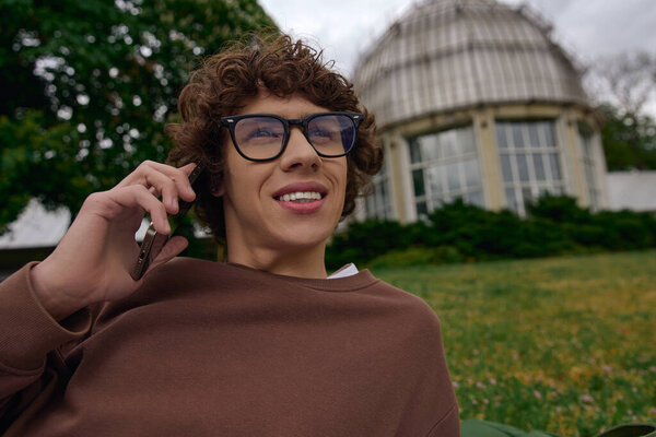 A handsome young man strolls through a lush landscape, engaged in a phone conversation with a smile.