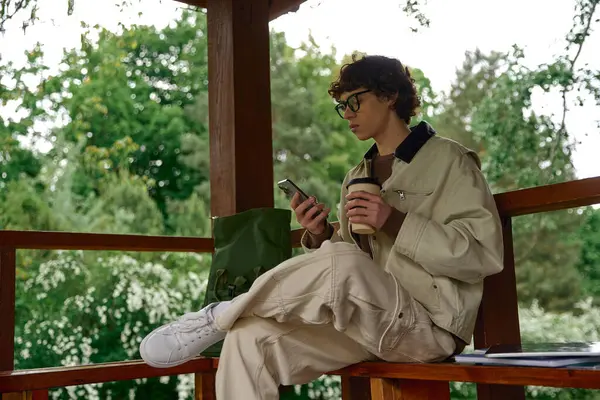 Genç adam dışarıda dinleniyor, kahvesini yudumluyor ve doğanın ortasında akıllı telefonuna dalmış..