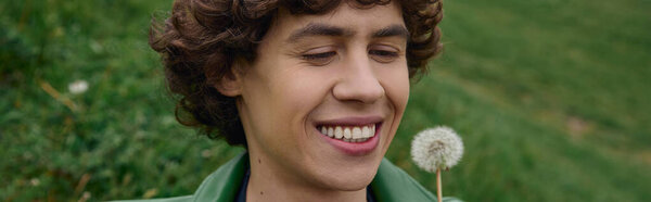 Handsome young man takes a serene walk outside, smiling at a delicate dandelion flower.