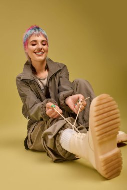 Kısa renkli saçlı genç bir kadın parlak bir stüdyoda otururken botlarını bağlıyor..