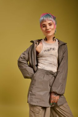 Neşeli genç bir kadın, kendine güvenen bir şekilde bir stüdyoda poz veriyor, canlı boyalı saçları ve şık kıyafetleri var..