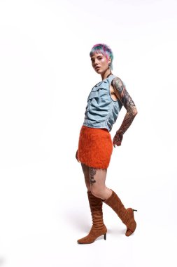 Genç bir kadın kendine güvenerek stüdyoda dikiliyor. Göz alıcı bir kıyafet ve renkli saçlar giyiyor..