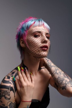 Genç bir kadın zarif bir şekilde stüdyoda poz veriyor, canlı saçlarını ve etkileyici dövmelerini sergiliyor..