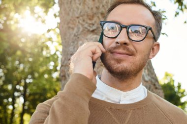 Genç bir adam, bir üniversite öğretmeni, telefonda konuşurken güneşli bir günün tadını çıkarıyor, gülümsüyor..