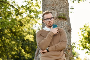Genç bir erkek öğretmen, elinde telefonla bir ağacın yanında duruyor ve huzurlu bir ortamda dalgın dalgın bakıyor..