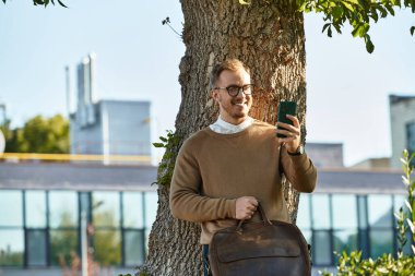 Kazaklı genç bir adam kampüsteki bir ağacın yanında telefonuna gülümsüyor..