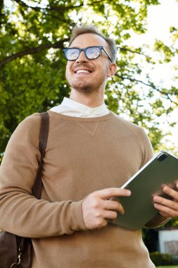 Genç adam yeşil bir açık havada elinde tabletle mutlu bir şekilde poz veriyor..