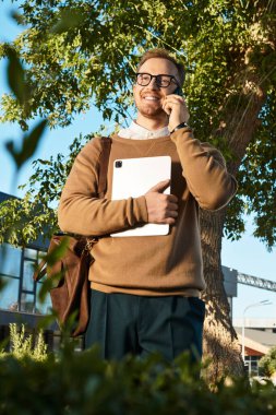 Genç bir adam, sıradan giyinmiş, elinde tabletle telefonda gülümsüyor ve dışarıda dikiliyor..
