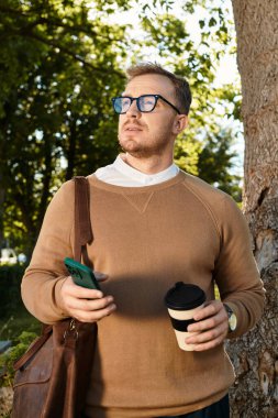 Sıradan giyinmiş genç bir adam, elinde kahve fincanı ve akıllı telefonuyla bir ağacın altında dikiliyor..