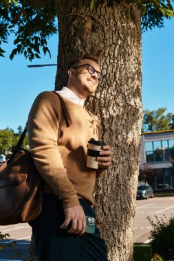 Bir adam ağacın yanında duruyor, gülümsüyor ve elinde bir kahve fincanı tutuyor, günlük giysiler içinde..