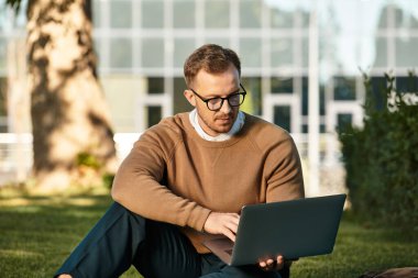 Genç bir erkek öğretmen çimlerin üzerinde oturur, dışarıda eğlenirken dizüstü bilgisayarda çalışır..