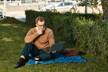 Genç bir erkek öğretmen yeşil bir alanda dizüstü bilgisayarıyla çalışıyor, açık havanın tadını çıkarıyor..