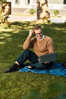 Genç bir üniversite öğretmeni parktaki bir battaniyeye oturmuş, bilgisayarını kullanarak mutlu mesut oturuyor..