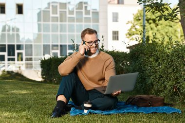 Neşeli genç bir öğretmen çim üzerinde otururken bir dizüstü bilgisayarla telefon kullanarak işe girişiyor..
