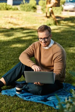 Genç bir adam parkta bir battaniyeye oturmuş, dizüstü bilgisayarına odaklanarak gülümsüyor..