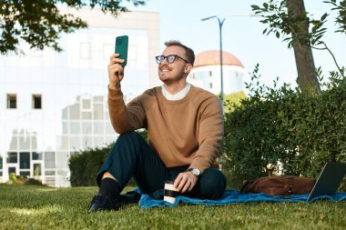 Genç bir adam çimlerin üzerinde oturur ve üniversite atmosferiyle çevrili bir şekilde selfie çekerken gülümser..