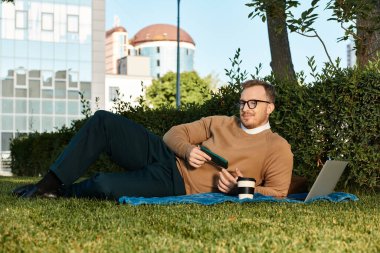 Delikanlı dışarıda çimde otururken dizüstü bilgisayar ve kahveyle rahatlamanın tadını çıkarıyor..