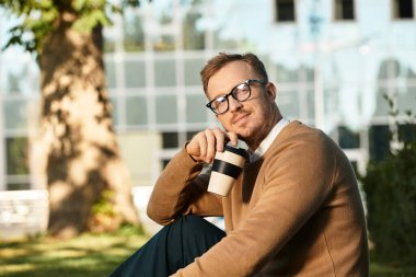Sıradan giyinmiş genç bir adam, üniversitenin dışında oturuyor, poz verirken kahvesinin tadını çıkarıyor..