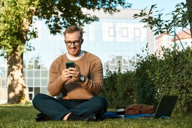 Genç bir adam, bir üniversite öğretmeni, açık havada akıllı telefonunu kullanarak çimlerin üzerinde oturuyor..