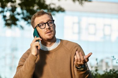 Genç bir adam, bir üniversite öğretmeni, açık hava ortamında el kol hareketi yaparken telefonuyla konuşur..