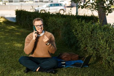 Genç bir adam elinde telefonla çimlerin üzerinde oturuyor, dışarıda konuşurken gülümsüyor..