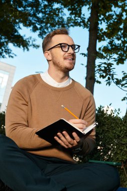 Genç öğretmen dışarıda oturur, gülümserken mavi gökyüzünün altındaki not defterine not alır..