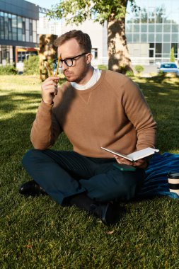 Genç bir erkek eğitimci çimlerin üzerinde oturur, elinde bir defter ve kalem, düşüncelerde kaybolur..