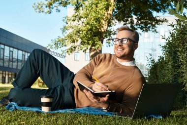Genç bir adam, gözlüklü, çimlerin üzerinde oturuyor, yakınlardaki bir dizüstü bilgisayara notlar yazıyor..