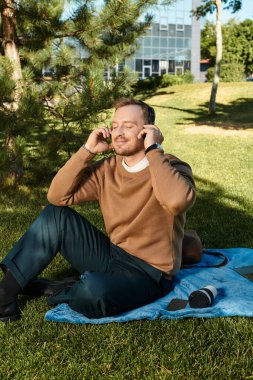 Genç bir adam parkta bir battaniyeye oturur, müzik dinler ve güneşin tadını çıkarır..