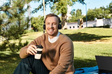 Genç bir erkek öğretmen parkta dinleniyor, kahvesini yudumluyor ve çalışırken müzik dinliyor..