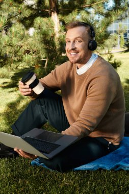 Genç bir adam çimlerin üzerinde oturuyor, dizüstü bilgisayarı ve kulaklıklarıyla çalışıyor ve elinde kahve fincanı var..