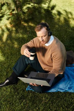Genç bir adam yeşil bir alanda mavi bir battaniyeye oturmuş laptopuyla çalışıyor ve kahvesini yudumluyor..