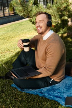 Genç bir adam yeşil bir alanda battaniyeye oturmuş elinde kahve fincanı tutarken gülümsüyor ve çalışıyor..