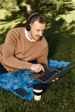 Üniversite öğretmeni parkta mavi bir battaniyenin üzerinde dinlenirken dizüstü bilgisayarıyla meşgul oluyor..