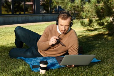 Genç bir erkek öğretmen, doğayla iç içe çimlerin üzerinde otururken dizüstü bilgisayarı üzerinde çalışıyor..