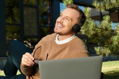 Genç bir üniversite öğretmeni laptopuyla çalışırken dışarıda rahatlıyor ve mutlu bir şekilde gülümsüyor..
