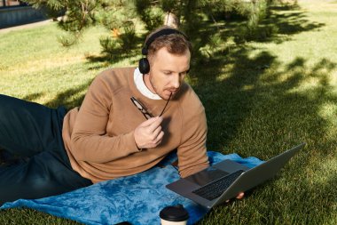 Rahat bir şekilde giyinen genç bir adam, yumuşak çimenlerde dinlenirken dizüstü bilgisayarıyla meşgul oluyor..