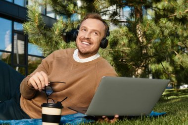 Genç eğitimci, dizüstü bilgisayarında kulaklıkla çalışırken ağaçların altında bir battaniyeye uzanıyor..