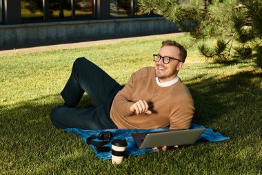Genç bir adam çimenlerde bir battaniyeye uzanıyor, dışarıda laptopuyla ve kahvesiyle ders veriyor..